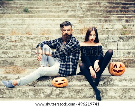 young halloween couple of bearded man with beard and mustache and girl in black tights sit on stony stairs with traditional autumn holiday symbol of orange spooky pumpkin outdoor