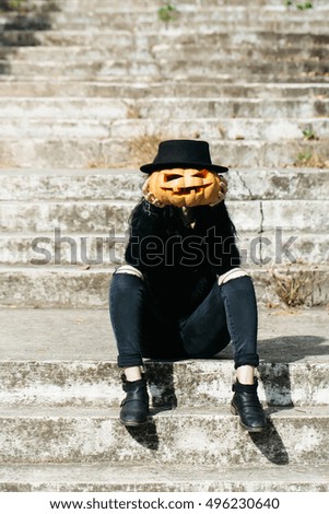 young halloween woman or girl in black witch hat and stylish jeans sitting on stony stairs with traditional autumn holiday symbol of orange spooky pumpkin outdoor