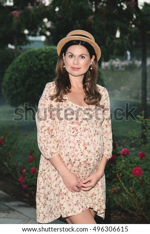 Beautiful pregnant girl waiting for a miracle. The fashionable photosession in park, a dress and a hat emphasizes refinement

