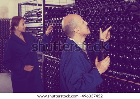 Positive mature man holding bottle of bubble wine and standing in winery production section