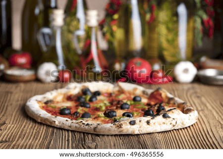 Delicious, tasty and fresh, rustic Italian pizza, served on wooden table.
Bunch of tomatoes, chili, basil, garlic in background. Bowls with different kinds of pepper. Many bottles of olive oil 