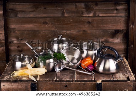 Different stainless steel pots, pans and laddles and fresh vegetables on old grunge wooden table against wood wall background