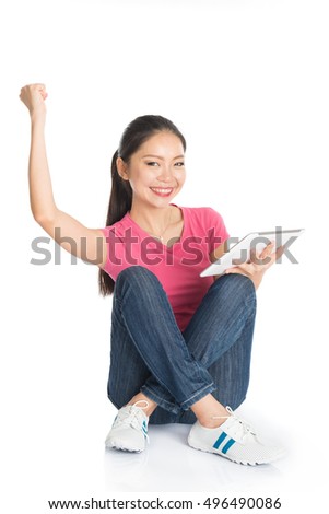 Full body young Asian girl in pink shirt using digital tablet computer arm raised celebrating success, seated on floor, full length isolated on white background.