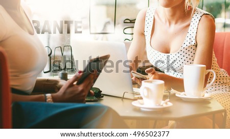 Young model look blonde woman is holding a smart-phone while sitting at the table with a laptop on it in a cafe. Two international female students are using electronicÂ� devices for writing a work.
