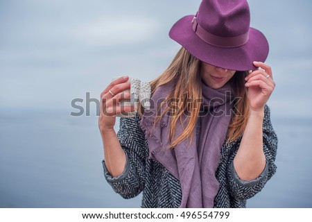 stylish girl in a burgundy hat stands on the background of the sea and holding a cup with angel wings