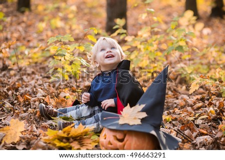 Portrait of Halloween smiling baby boy in dracula costume (cloak). Child in autumn forest looking at the falling leaves. Halloween pumpkin, witch hat, holiday concept