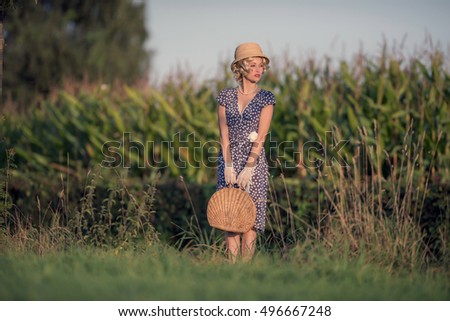 Vintage 1920s summer fashion woman with blue dress and straw hat standing with handbag in rural landscape.