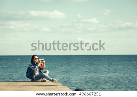 happy stylish woman hipster holding photo camera and smiling at sunny shore near water, summer travel concept, space for text