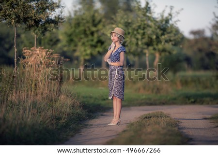 Retro 1920s summer fashion woman with blue dress and straw hat standing on rural road.