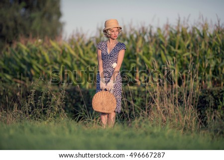 Vintage 1920s summer fashion woman with blue dress and straw hat standing with handbag in rural landscape.