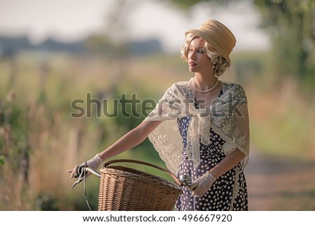 Retro woman dressed in 1930s fashion standing with bicycle in rural landscape.