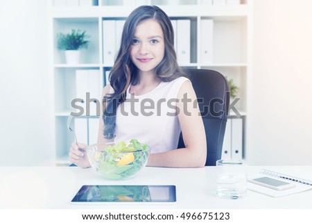 Business lady eating salad at her table at work in office. She is smiling and enjoying her lunch break. Concept of healthy lifestyle and dieting. Toned image