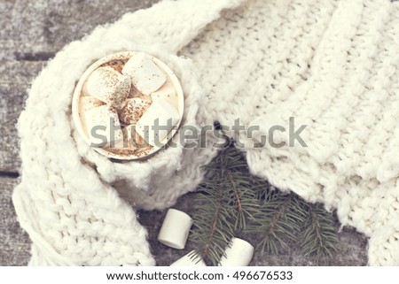 A cup of warm cocoa with marshmallows and a cozy knitted scarf on old wooden boards in the snow