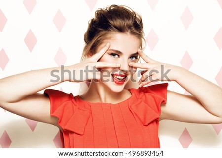 Portrait of young beautiful woman with light  eye makeup and red lipstick. Smiling. Touching her face. Wearing red dress. Pink rhombus background.