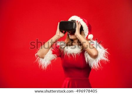 christmas, x-mas, winter, happiness concept - Beautiful young brunette with long hair in santa helper hat wearing virtual reality headset over red background