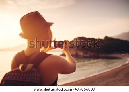 Woman traveler with smartphone taking photo near sea at sunset