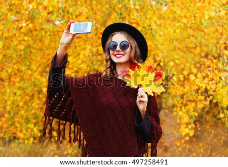Autumn portrait of stylish happy smiling young woman taking selfie with smartphone and yellow maple leaves wearing round hat, brown knitted poncho in the park 