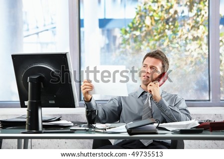 Smiling professional businessman on landline call looking at paper held in hand siting at office desk.