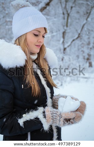 Young pretty girl in a winter coat and white hat standing in a snowy forest and holding hearts made of snow