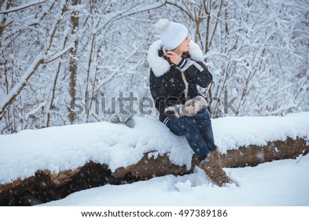 Young beautiful blonde girl sitting on the big snow-covered log in a snowy park. Snowy winter
