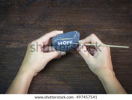 image of a woman's hand holding words "Hope" brush painted on river stone over wooden background, vignette and vintage tone, conceptual image