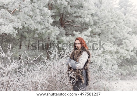 beautiful gentle girl with red hair in a fur vest standing in a snowy forest with iniem on the branches of trees