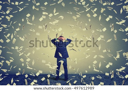 Rear back view of business man standing in front of a wall under money rain dollar banknotes falling down, hands on head wondering. Full body length of businessman facing the wall