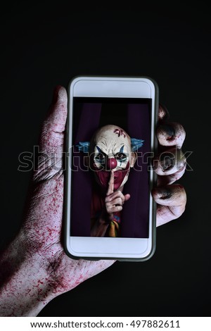 closeup of a scary and bloody hand holding a smartphone with a picture of a scary evil clown asking for silence in its screen