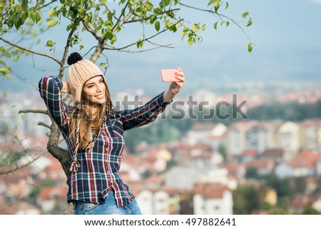 Millennial young blonde Caucasian woman taking a selfie on smart phone, outdoors on sunny autumn day. Teenage girl in checkered shirt, denim jeans and knitted beanie hat taking a photo of herself.