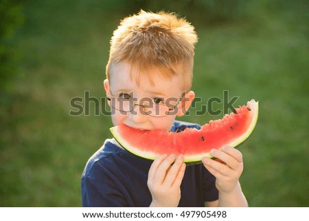Happy baby boy with red hair in blue shirt and eyeglasses on happy smiling face eating watermelon summer day outdoor on green natural background