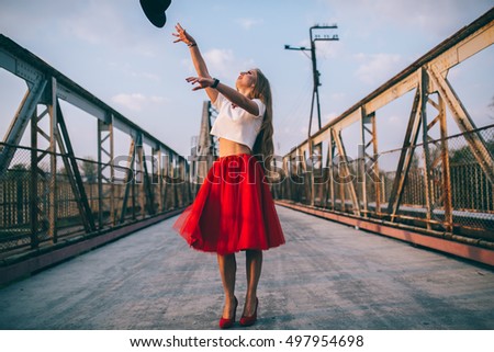 Outdoor lifestyle portrait of happy blonde young woman in stylish casual red dress (skirt) and black hat stay on bridge on the street. Girl throws hat and jump on the bridge. Having fun . Long hair