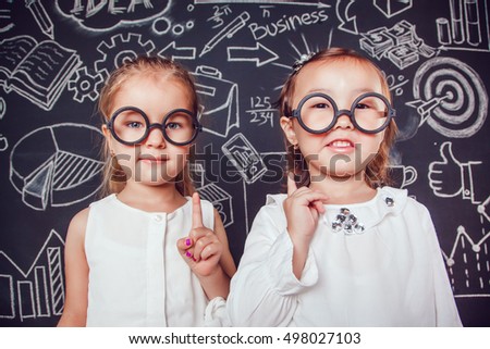 Two little smart girls in glasses lifting finger up on a background of wall with business or school picture
