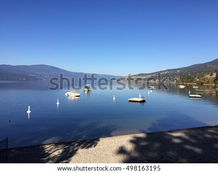 Calm summer lake with bright clear blue sky over clear blue lake water. Boats anchored and buoys on water. Mountains in background and reflections on water.  