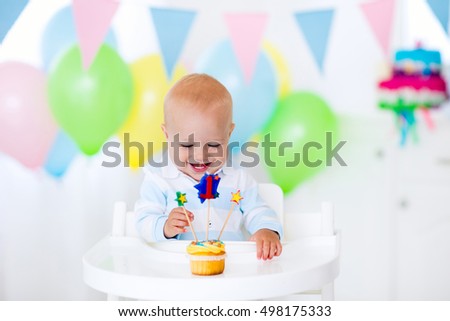 Adorable baby boy celebrating first birthday blowing candles on colorful cup cake. Kids birthday party decorated with balloons and pastel color banner. Child eating cake and candy.