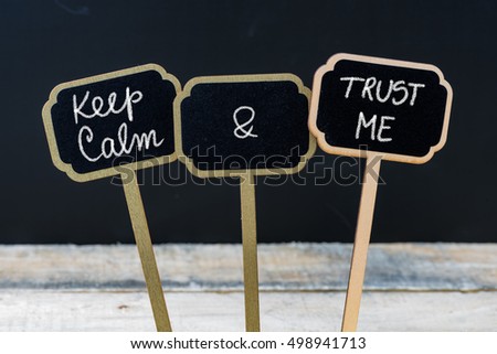 Keep Calm and Trust Me message written with chalk on mini blackboard labels, defocused chalkboard and wooden table in background. Fun and humor concept