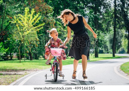 Beautiful and happy young mother teaching her cute daughter to ride a bicycle. Both smiling and looking at each other. Summer park in background Royalty-Free Stock Photo #499166185