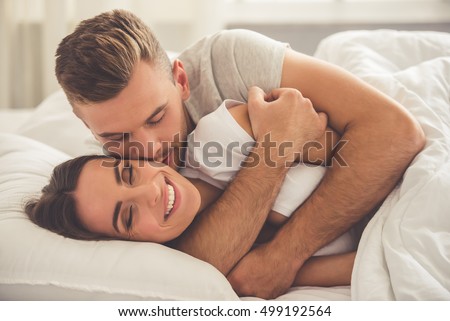 Handsome young man is hugging and kissing his beautiful smiling wife in cheek while they both are lying in bed in the morning Royalty-Free Stock Photo #499192564