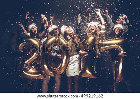 New 2017 Year is coming! Group of cheerful young people in Santa hats carrying gold colored numbers and throwing confetti  Royalty-Free Stock Photo #499219162