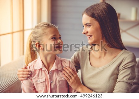 Portrait of beautiful young mother and her little daughter hugging, looking at each other and smiling while sitting on sofa at home