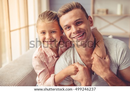 Portrait of handsome young father and his little daughter hugging, looking at camera and smiling while sitting on sofa at home Royalty-Free Stock Photo #499246237