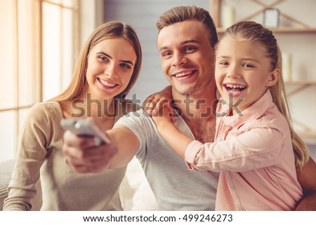 Beautiful happy young parents and their little daughter watching TV and smiling while sitting on sofa at home