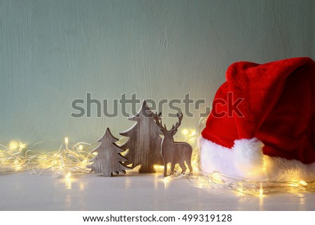 Low key Image of wooden christmas tree and deer with garland warm lights next to santa hat
