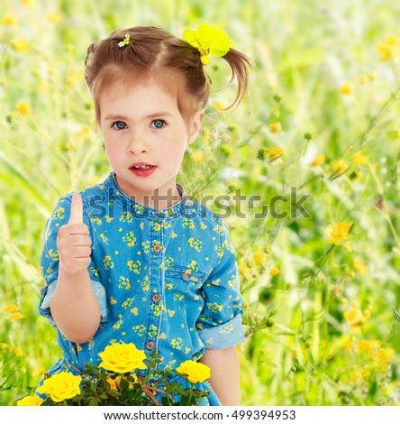 Cute little girl in a blue dress. The girl shows the thumb of the right hand. Before her a bouquet of yellow flowers.On blurred background of green grass with yellow flowers