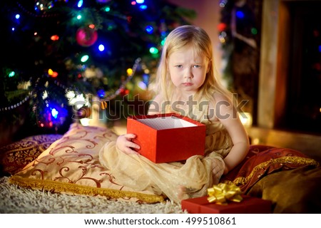 Cute little girl is unhappy with her Christmas gift by a fireplace in a cozy dark living room on Xmas eve