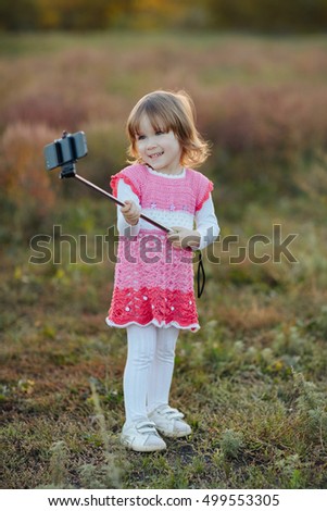 happy little girl taking selfie-stick with smartphone, monopod in autumn park and photographing themselves smiling on background autumn forest. family, childhood, season, technology, people concept 