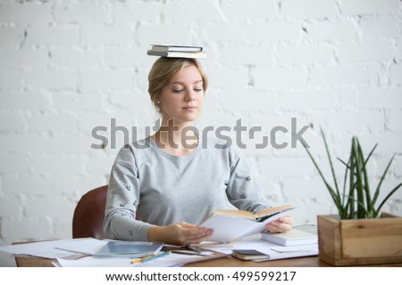 Portrait of a young attractive woman at the desk with books on her head, sitting straight, reading a book. Education concept photo, lifestyle Royalty-Free Stock Photo #499599217
