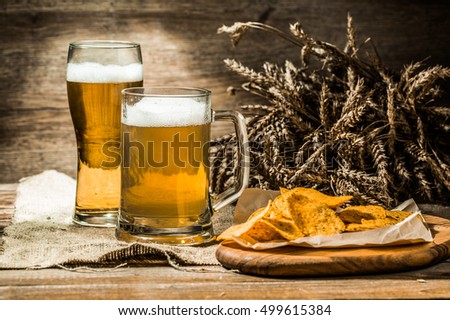 Mug, glass of foamy beer on wooden table with wheat spikelets and potato chips on board