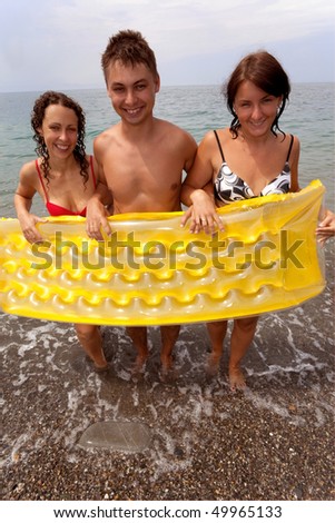 trio of young people stand on seacoast and hold yellow inflatable mattress. youth group has rest on beach