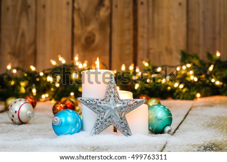Christmas background with white candles, holiday ornaments, metal star and string of lights with green garland border in snow; red, teal blue, silver and gold rustic Christmas background