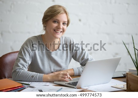 Portrait of a young smiling student girl sitting at the desk working with a laptop, education concept photo, looking at the camera